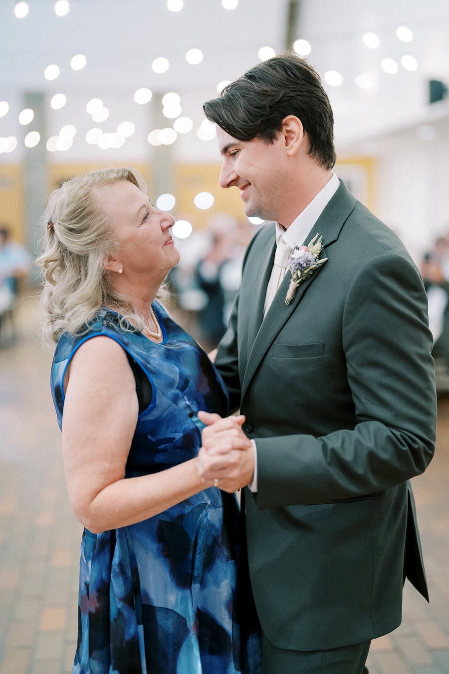 A man in a suit dances with a woman in a blue dress at an indoor event, surrounded by soft, glowing lights.