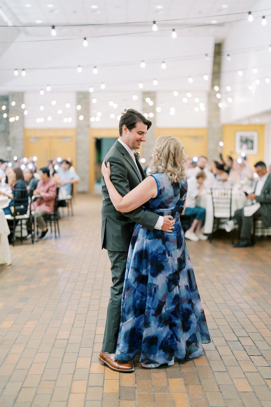A couple dancing together at a wedding reception, surrounded by elegantly dressed guests under softly glowing string lights in a warmly lit venue.