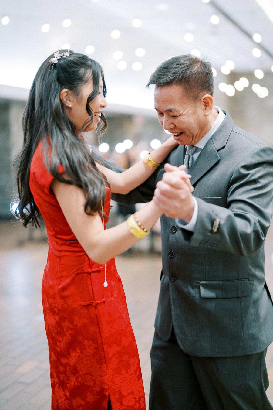 Father and daughter joyfully dancing at a wedding reception, with the daughter in a vibrant red dress and the father in a gray suit, under twinkling lights.