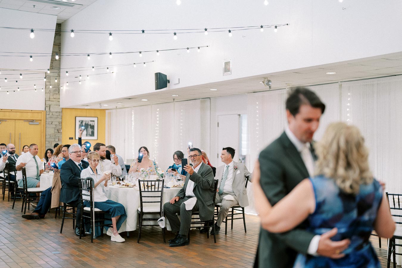 A couple dances in a warmly lit reception hall while guests seated at tables watch and capture the moment on their phones, surrounded by elegant string lights.