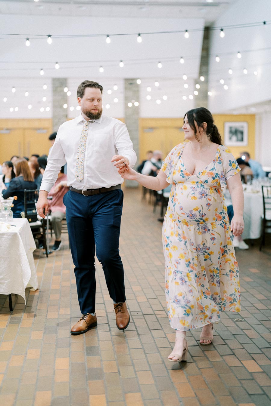 A couple walking hand-in-hand at a formal event, with the man wearing a white shirt and blue pants and the woman in a floral dress, under a canopy of string lights.