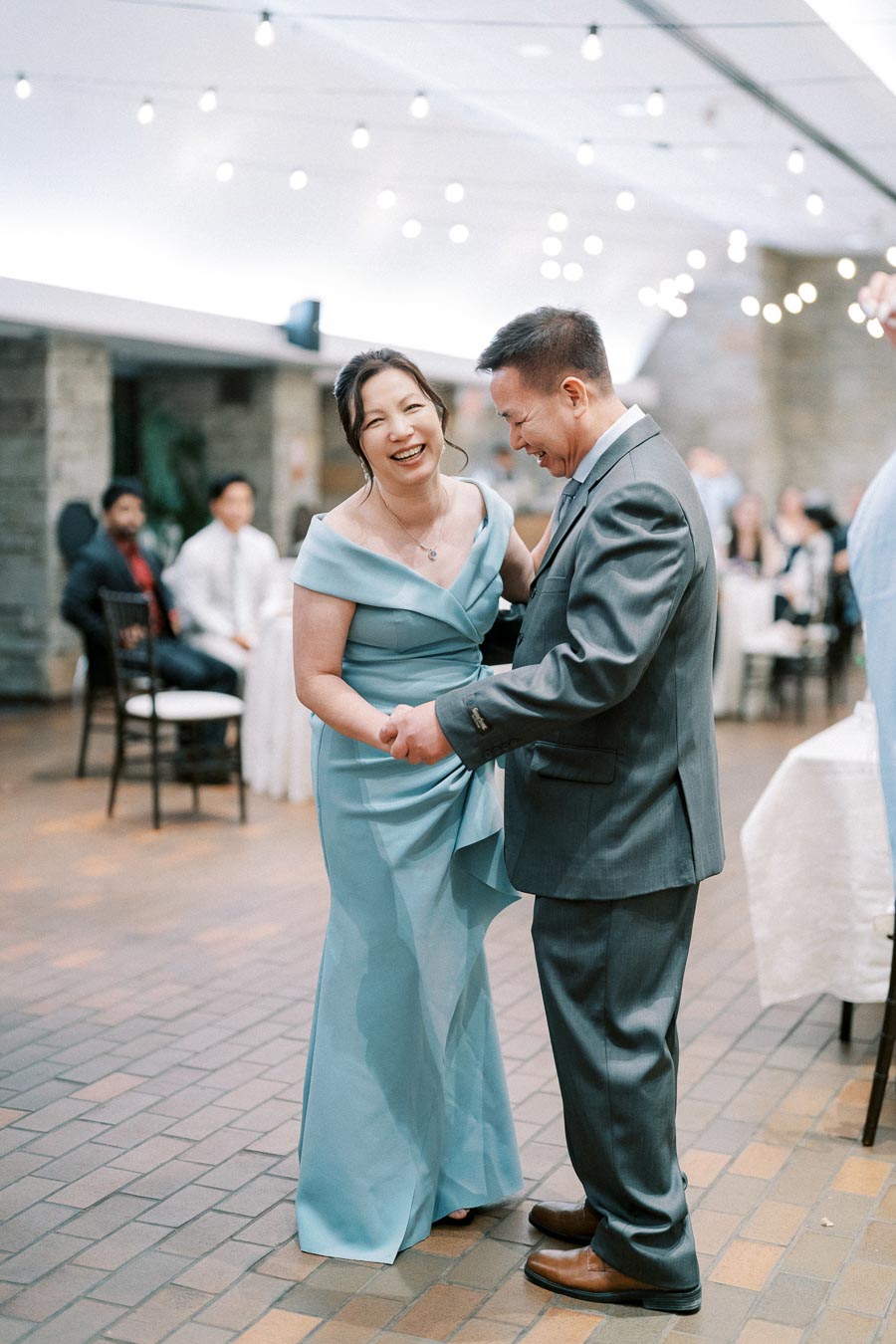 A couple dressed in formal attire joyfully dancing at a wedding reception, with decorative string lights in the background.