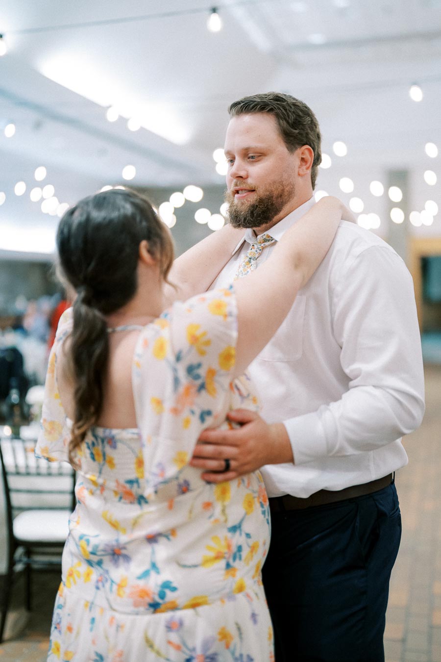 A couple dancing closely at a wedding reception, under soft string lights, dressed in a white shirt and floral dress, creating a romantic atmosphere.