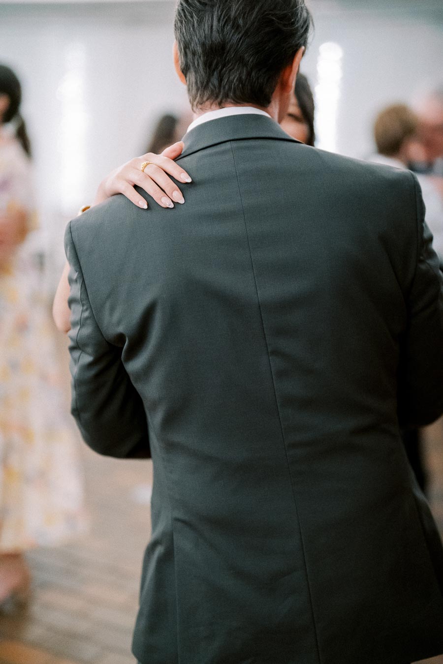 A couple embracing and dancing at a formal event, with the focus on the back of a man in a suit and a woman's hand resting on his shoulder.