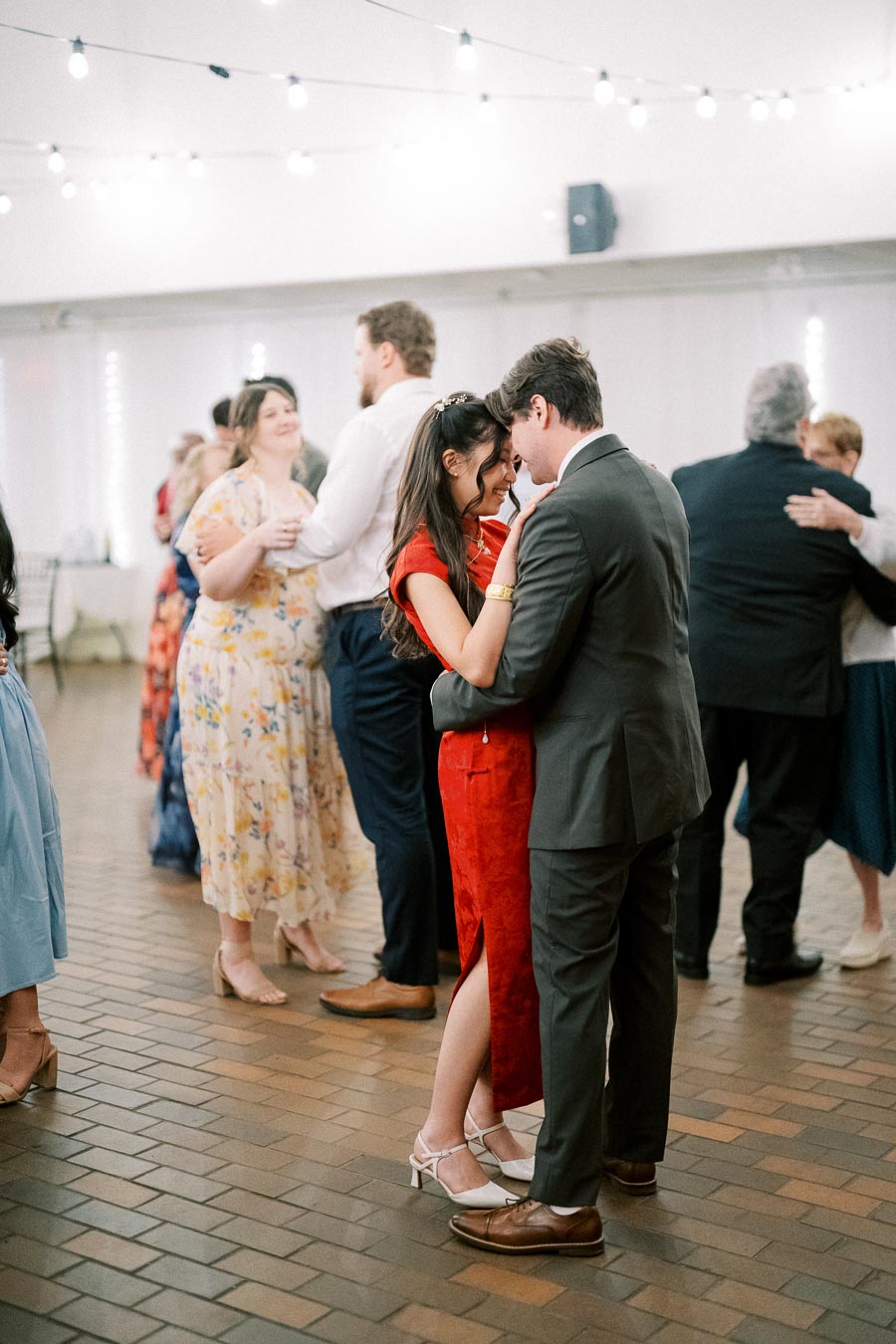 A couple enjoys a romantic dance at a wedding reception in a warmly lit venue, surrounded by other dancing guests.