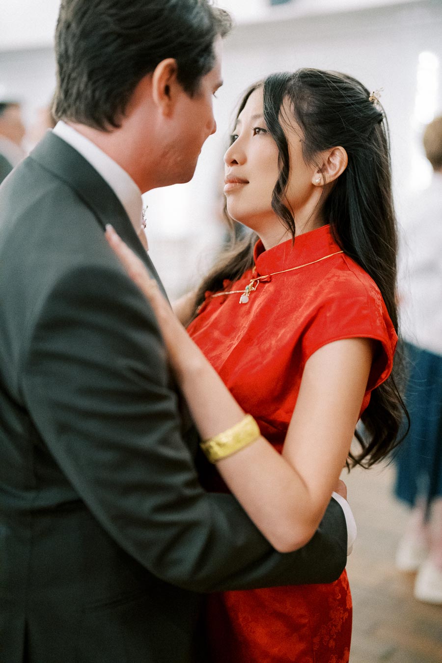 A couple intimately embracing at a special event, with the woman wearing a traditional red dress and gold bracelet, and the man in a suit, highlighting cultural celebration and romantic connection.