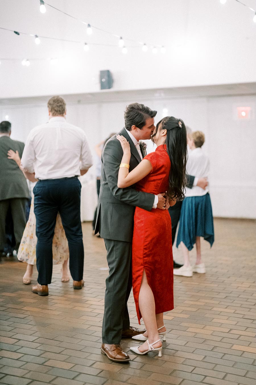 A couple dancing and kissing at a wedding reception, with the woman wearing a red dress and the man in a suit, surrounded by other dancing guests under string lights.