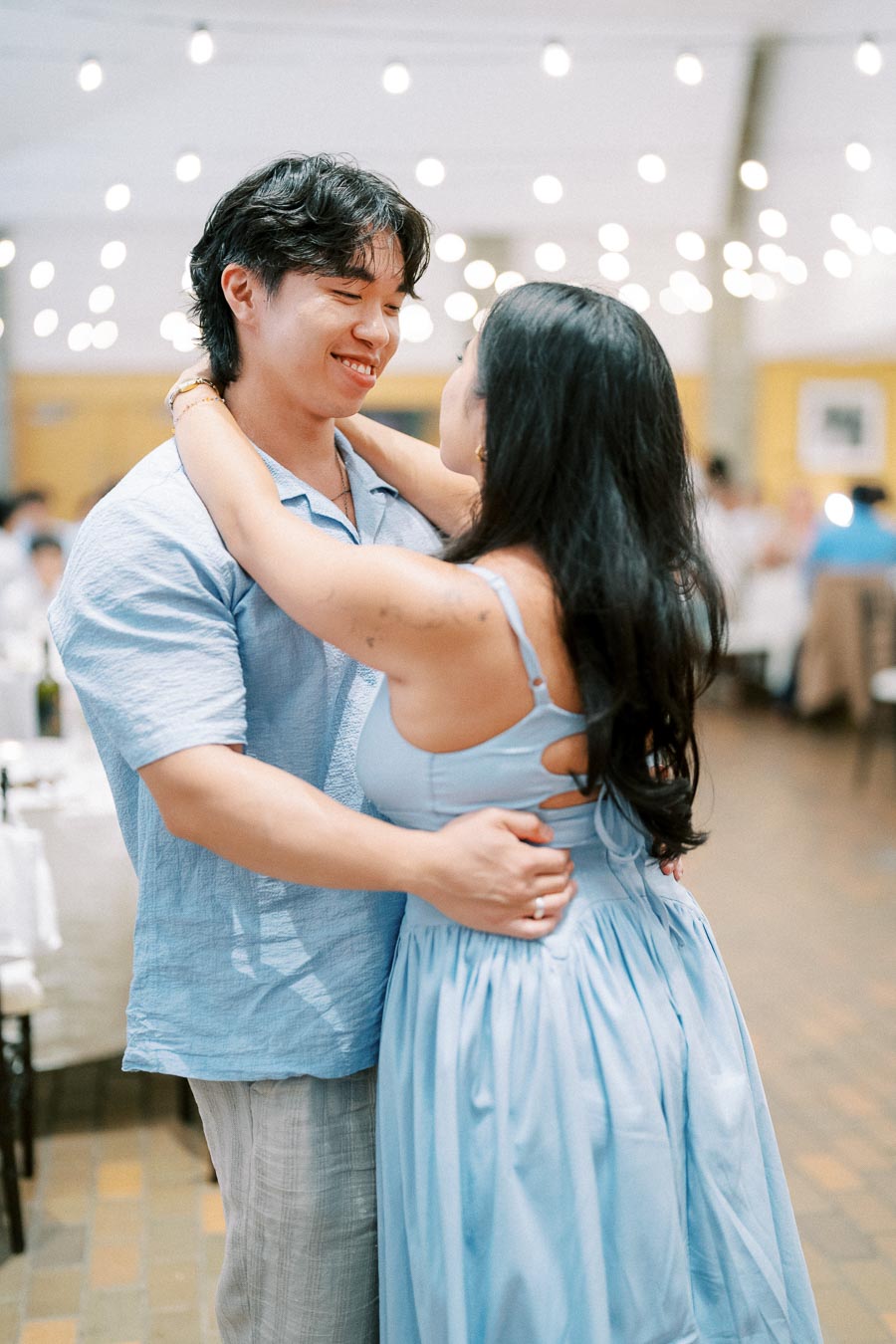 Couple in matching blue outfits dancing at an elegant indoor event with soft lighting and hanging string lights.