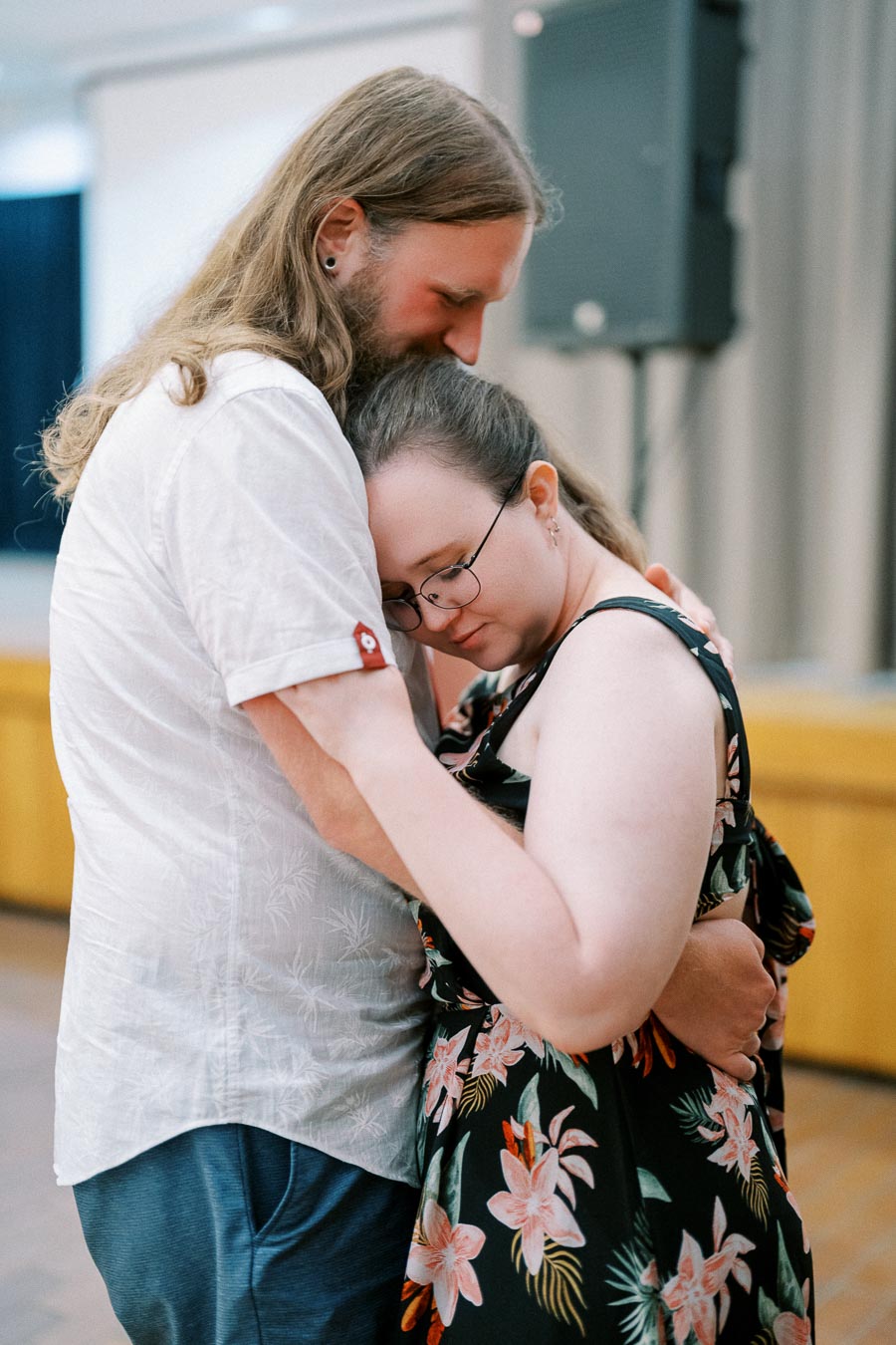 A couple sharing an intimate hug indoors, with the woman resting her head on the man's shoulder, both expressing tenderness and affection in a warm setting.
