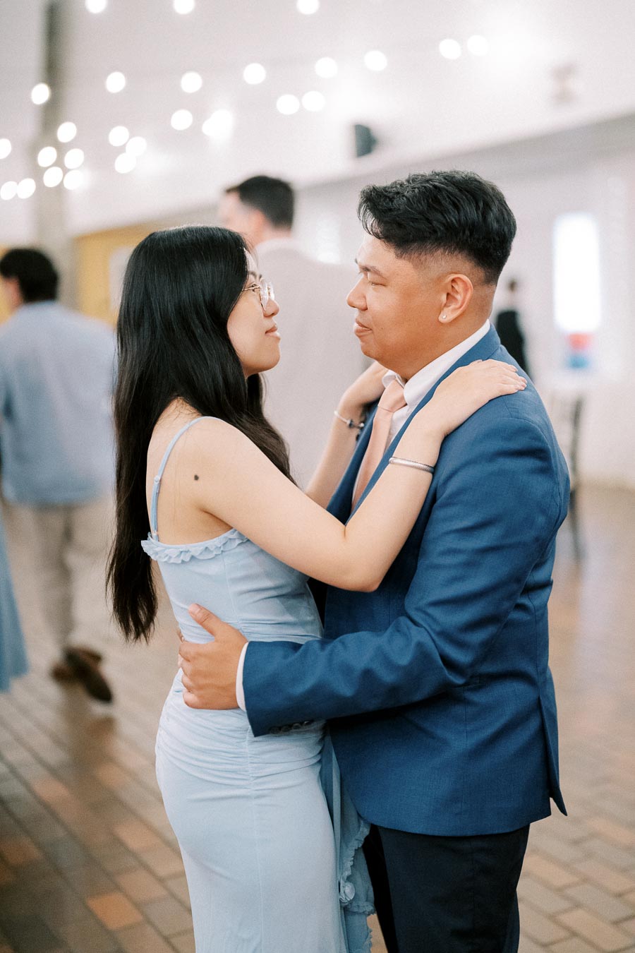 A couple dancing closely at a formal event, both wearing matching blue outfits, with soft lighting and blurred people in the background, creating a romantic atmosphere.