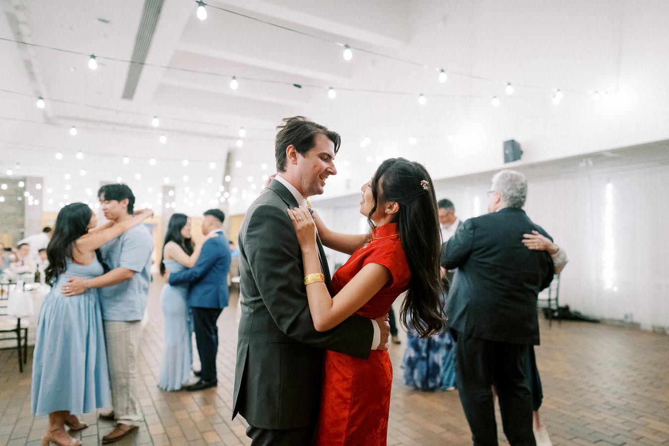 A couple in formal attire dances under string lights at a wedding reception, surrounded by other guests on the dance floor.