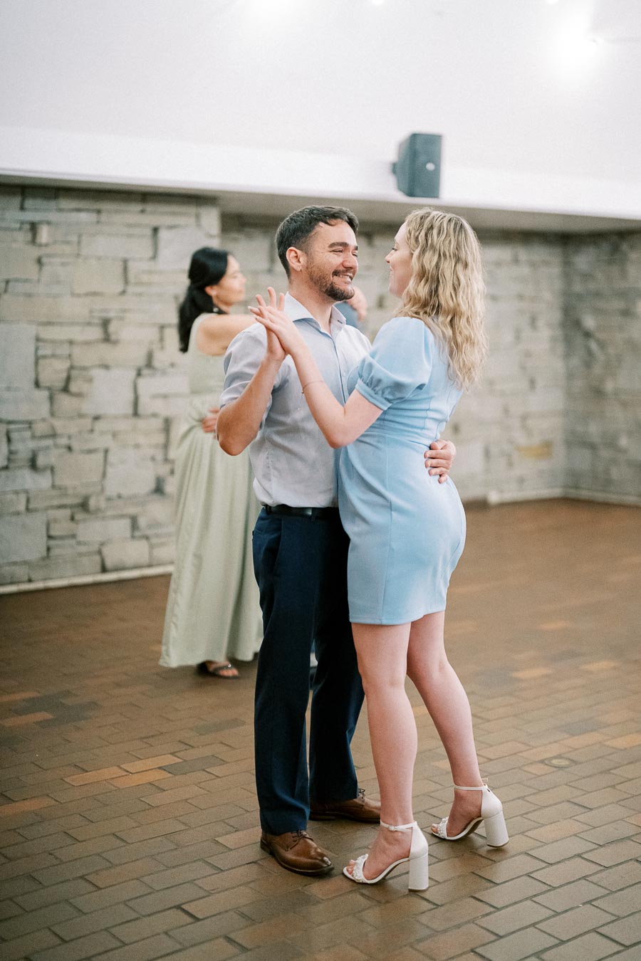 A couple dancing closely at a wedding reception, with a woman wearing a light blue dress and a man in a light shirt and dark pants. The background shows another couple dancing and a stone wall, creating a romantic atmosphere.