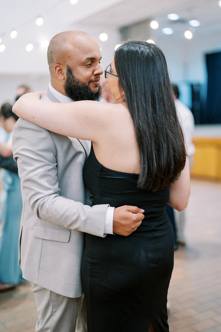 A couple sharing a romantic dance at a wedding reception, with string lights in the background creating an intimate atmosphere. The man is wearing a light gray suit, and the woman is in a black dress.