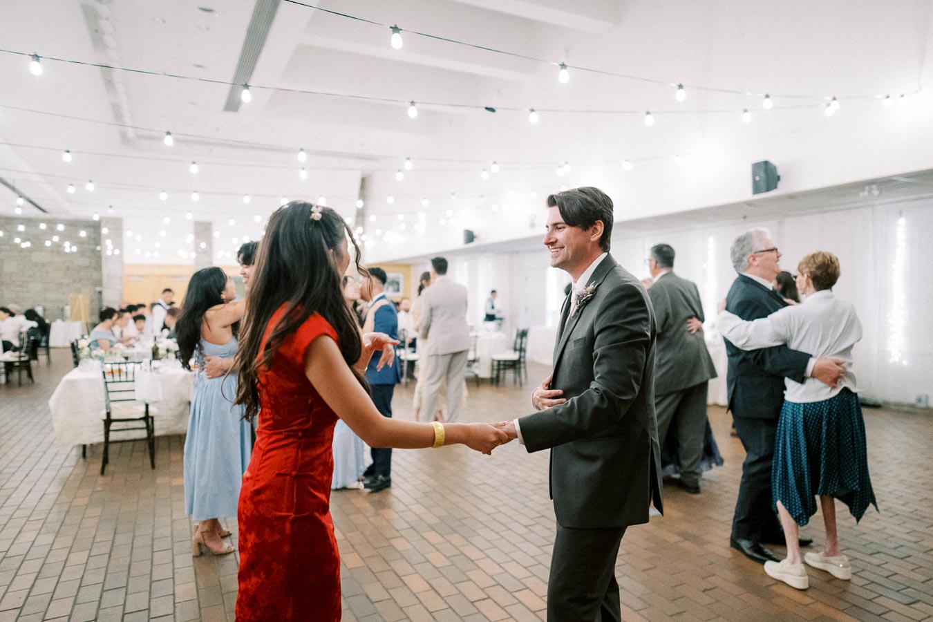 A lively dance floor at a wedding reception, featuring elegantly dressed guests, including a woman in a red dress and a man in a suit, enjoying a festive atmosphere with string lights hanging above.
