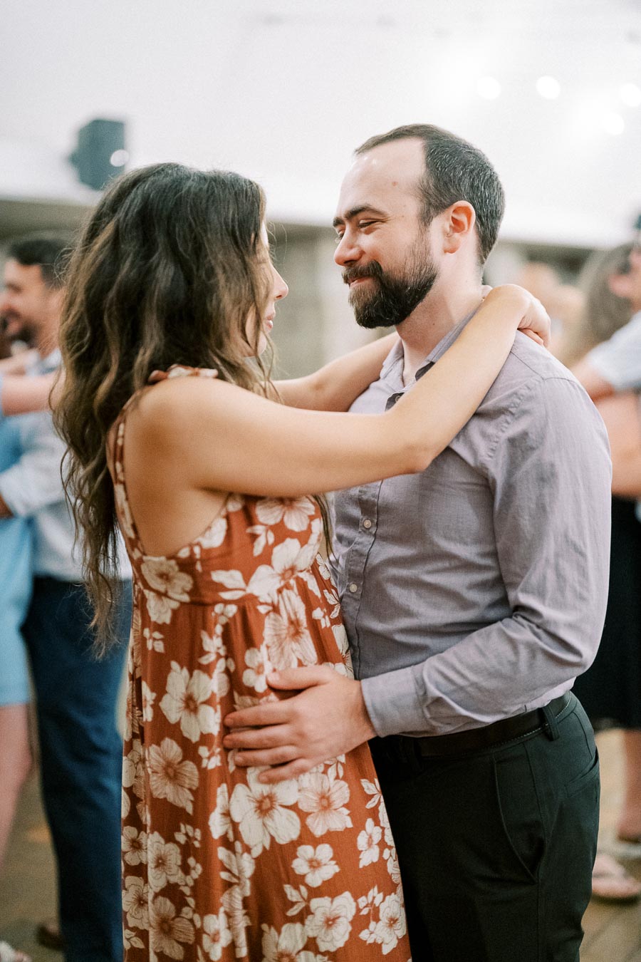 Couple dancing closely at a wedding reception, the woman in a floral dress and the man in a gray shirt, sharing a joyful moment surrounded by other guests.
