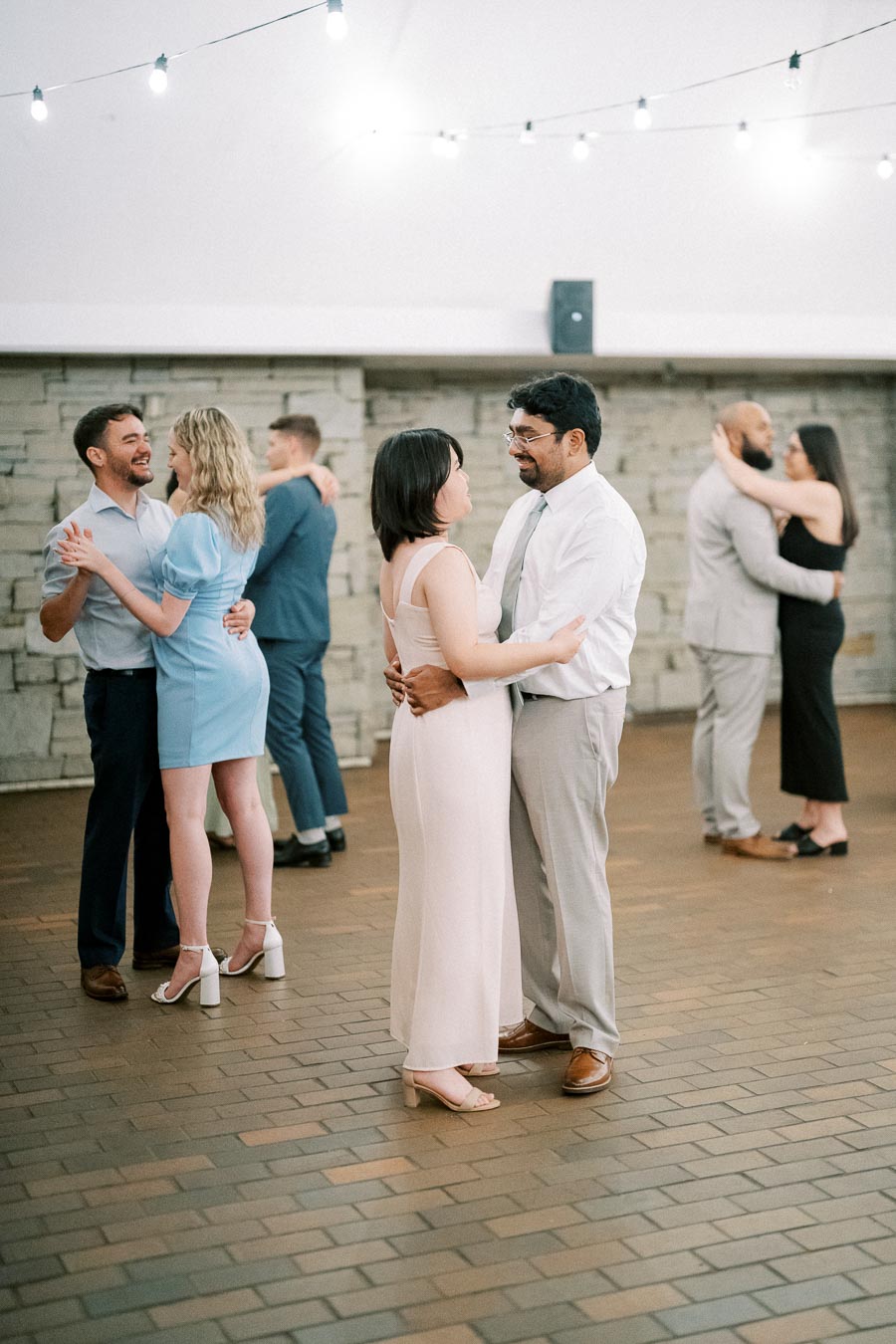 Couples dancing at an indoor event with string lights and stone wall backdrop, showcasing a joyful and elegant atmosphere.