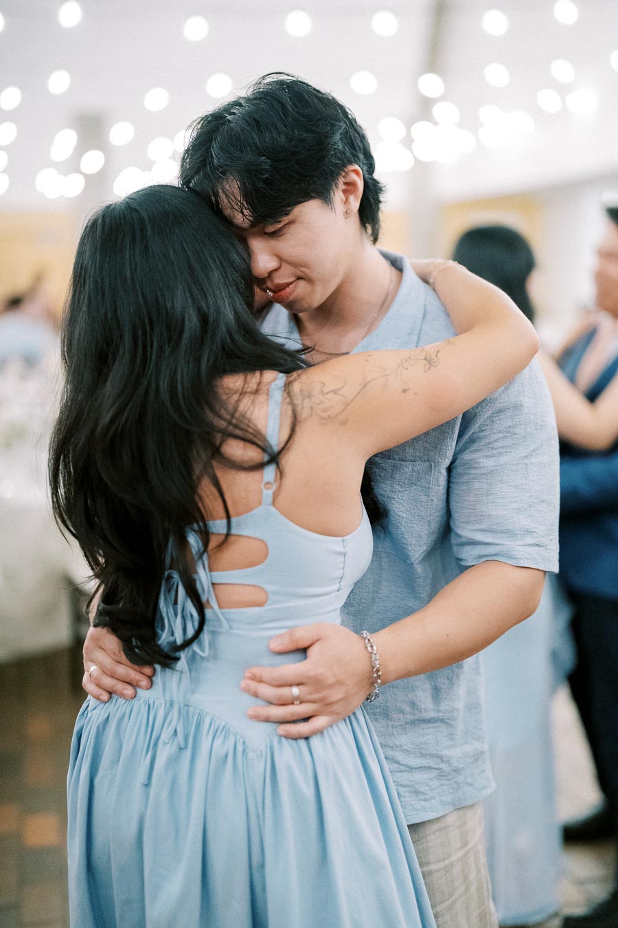 A couple lovingly embraces and dances at a celebration, with soft lighting and an elegant atmosphere, both wearing blue clothing.