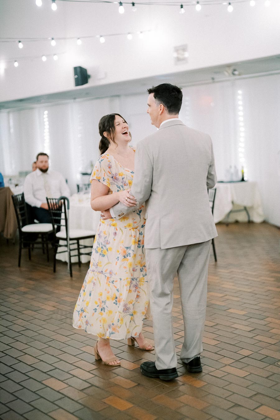 A couple dancing and laughing at a wedding reception, with string lights above and guests seated in the background.