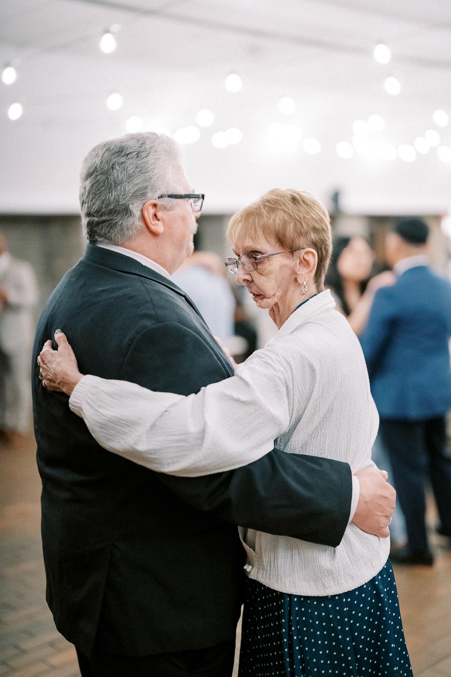 Elderly couple dancing at an event under string lights, capturing a tender moment with other guests in the background.