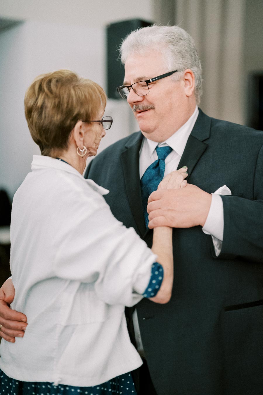 Elderly couple sharing a tender moment while dancing at an event, wearing formal attire and holding hands lovingly.