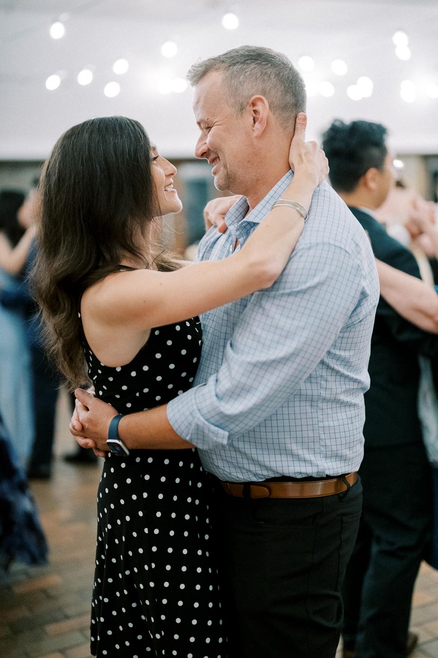 A couple dancing closely at an indoor event, illuminated by string lights, creating a romantic ambiance. The woman is wearing a black polka dot dress, and the man is in a checkered shirt.