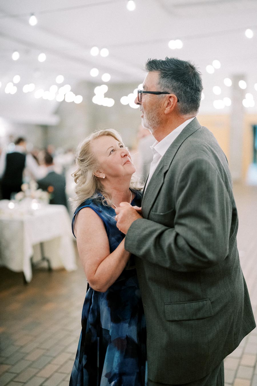 Older couple dancing romantically at an elegantly lit wedding reception, with string lights in the background creating a warm, festive atmosphere.