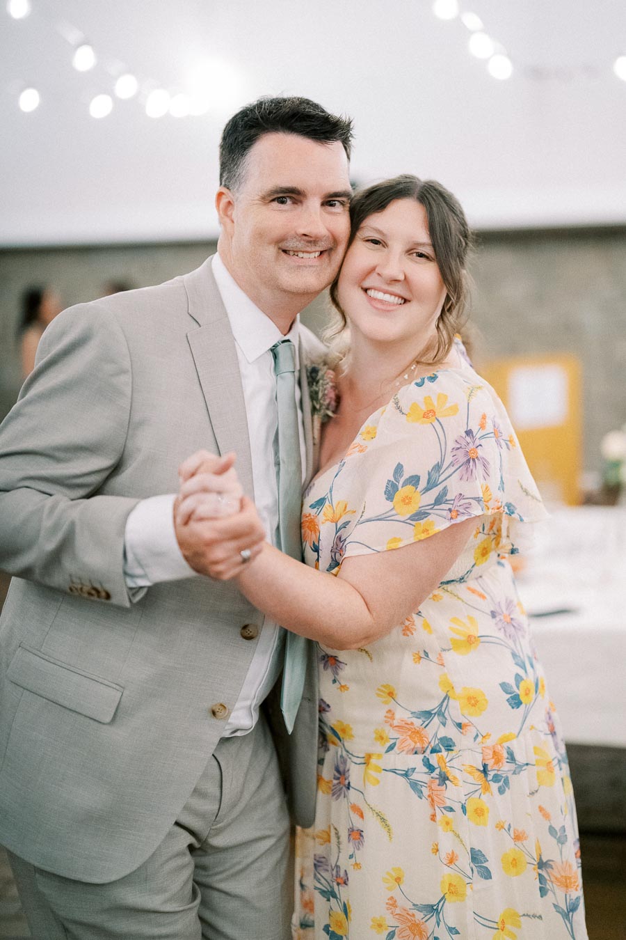 A smiling couple dancing closely at a wedding reception, with string lights and soft decorations in the background, creating a romantic ambiance. The woman is wearing a floral dress, and the man is in a light gray suit.