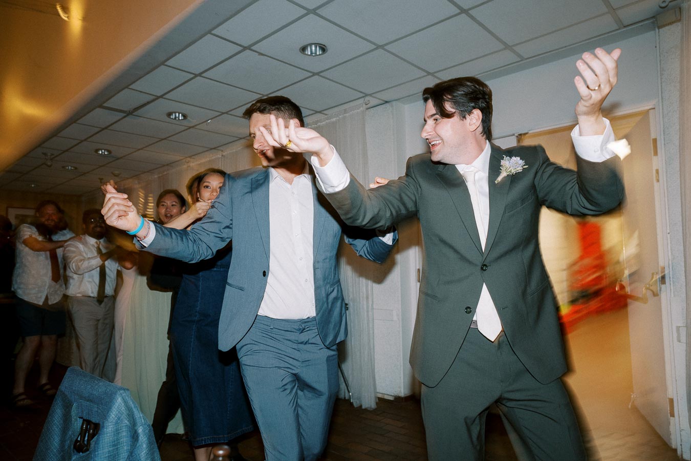 A group of people joyfully dancing in a line at a wedding reception, with the man in front wearing a green suit and white shirt, followed by others in formal attire. The atmosphere is lively and festive, capturing a moment of celebration.