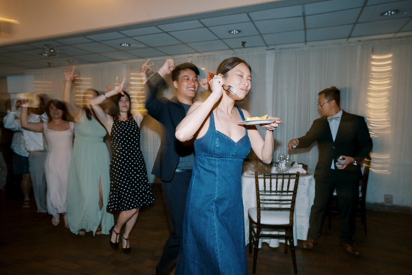 People joyfully dancing in a conga line at a lively indoor celebration, with a woman leading while holding a plate of food.