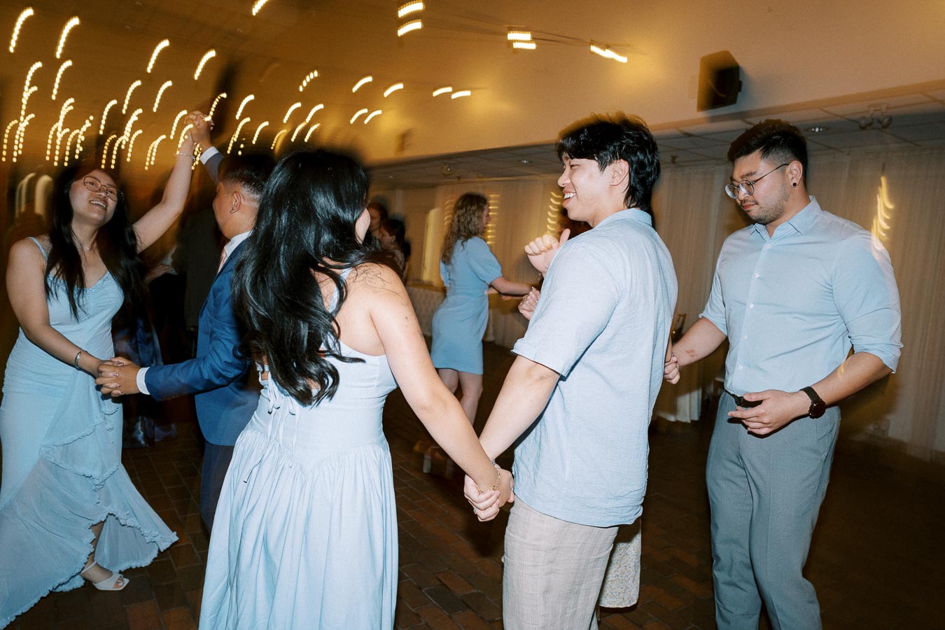 A group of people in blue outfits dancing joyfully at an indoor party or wedding reception, with soft lighting and blurred motion capturing the dynamic atmosphere.