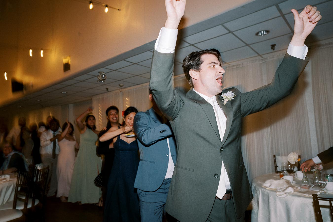 Man in a suit leading a conga line at a lively wedding reception, with guests joyfully dancing in a decorated banquet hall.
