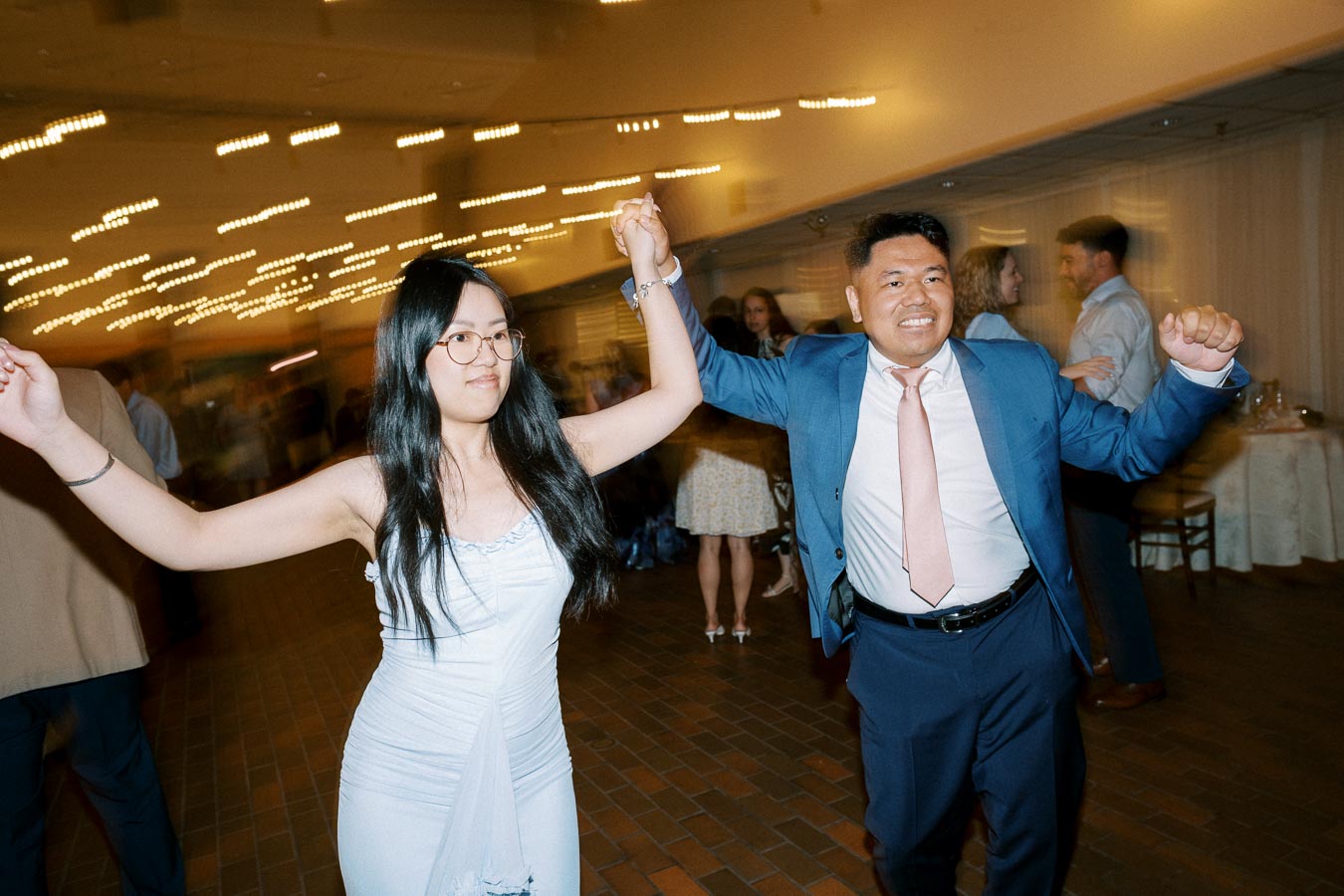 A man in a blue suit and a woman in a white dress dance joyfully at a lively indoor event, with blurred lights and guests in the background.