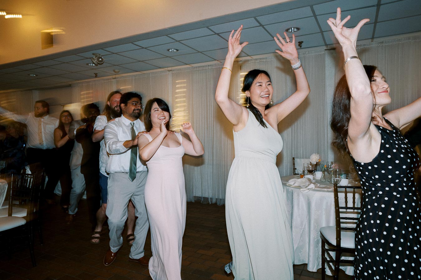 A lively group of people enjoying a conga line at a wedding reception, with joyful expressions and festive attire, creating a fun and celebratory atmosphere in an event hall.