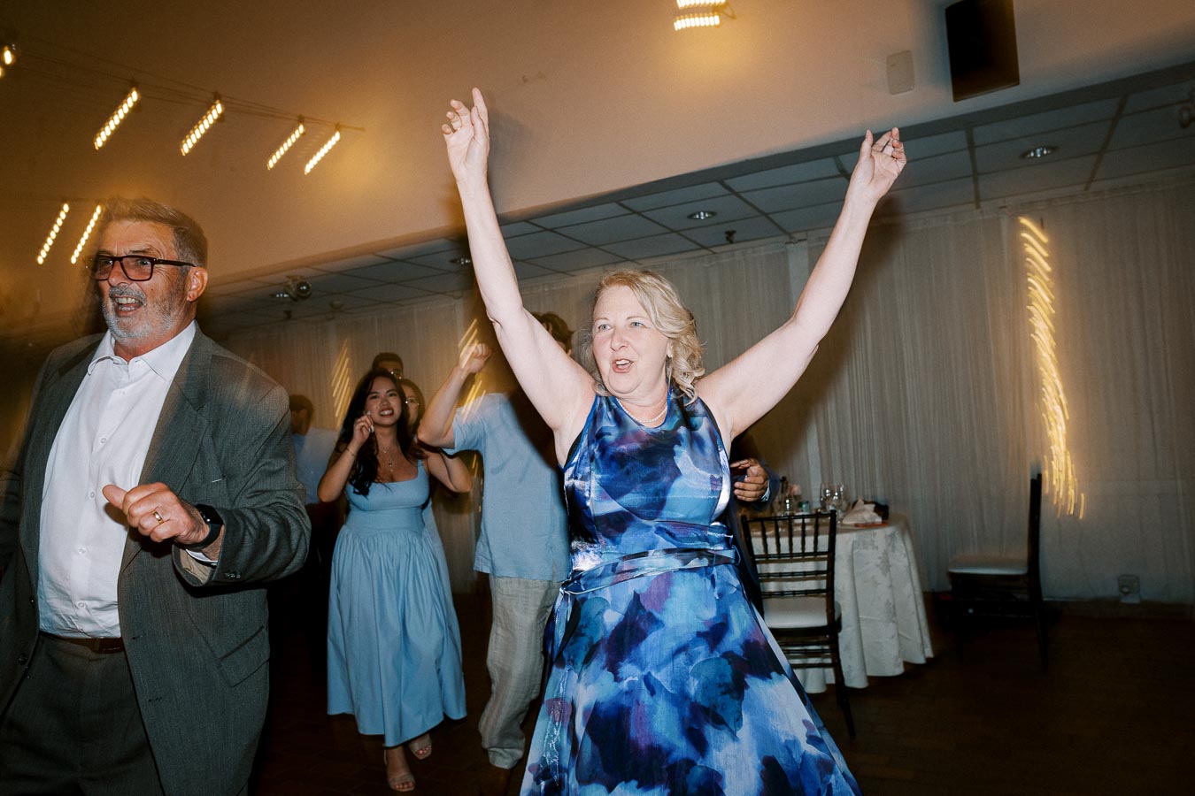 A joyful group of people dancing at a lively indoor event, with women in elegant blue dresses and a man in a suit, celebrating in a warmly lit venue.