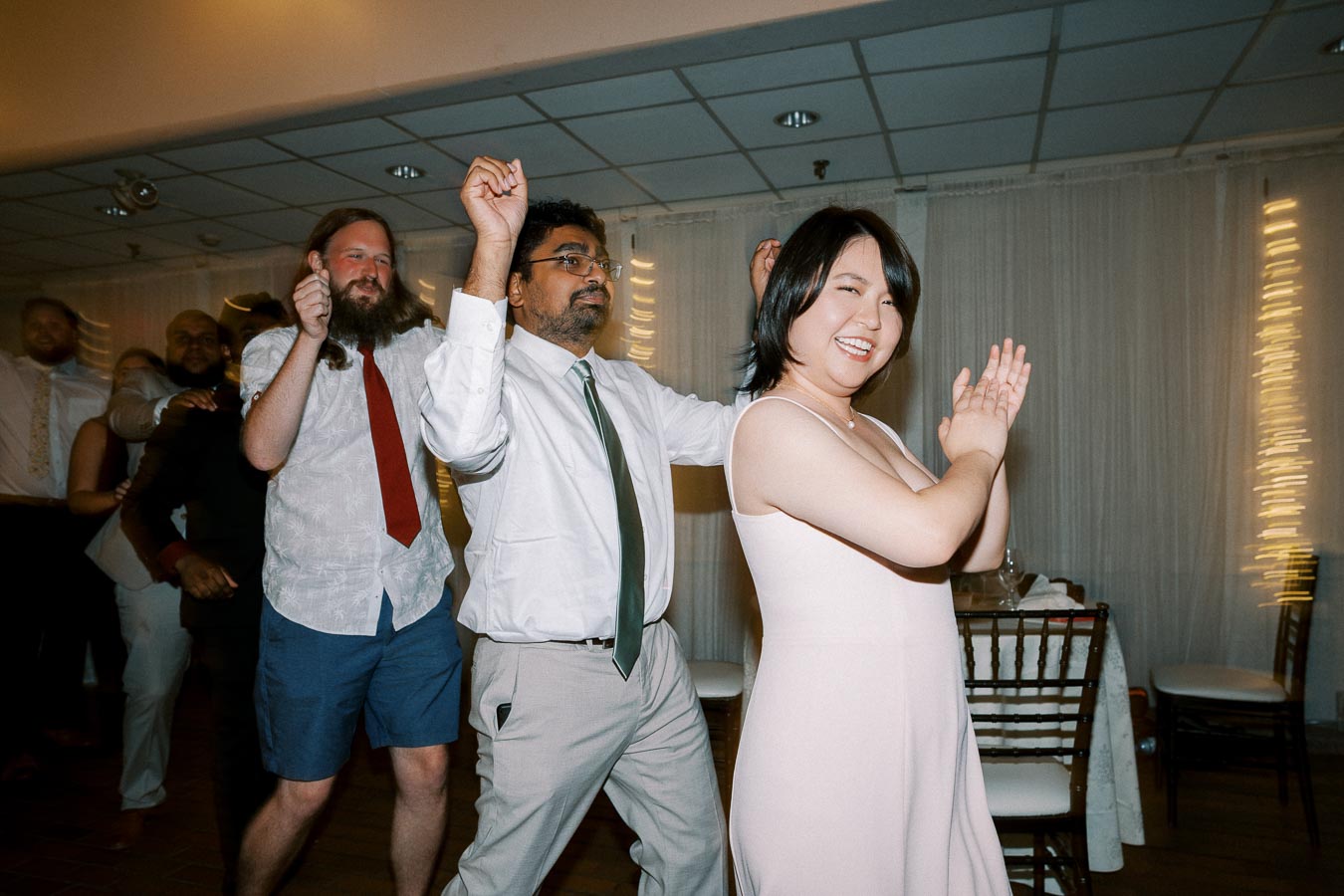 A group of people joyfully dancing in a line at a festive indoor event, with string lights creating a warm ambiance.