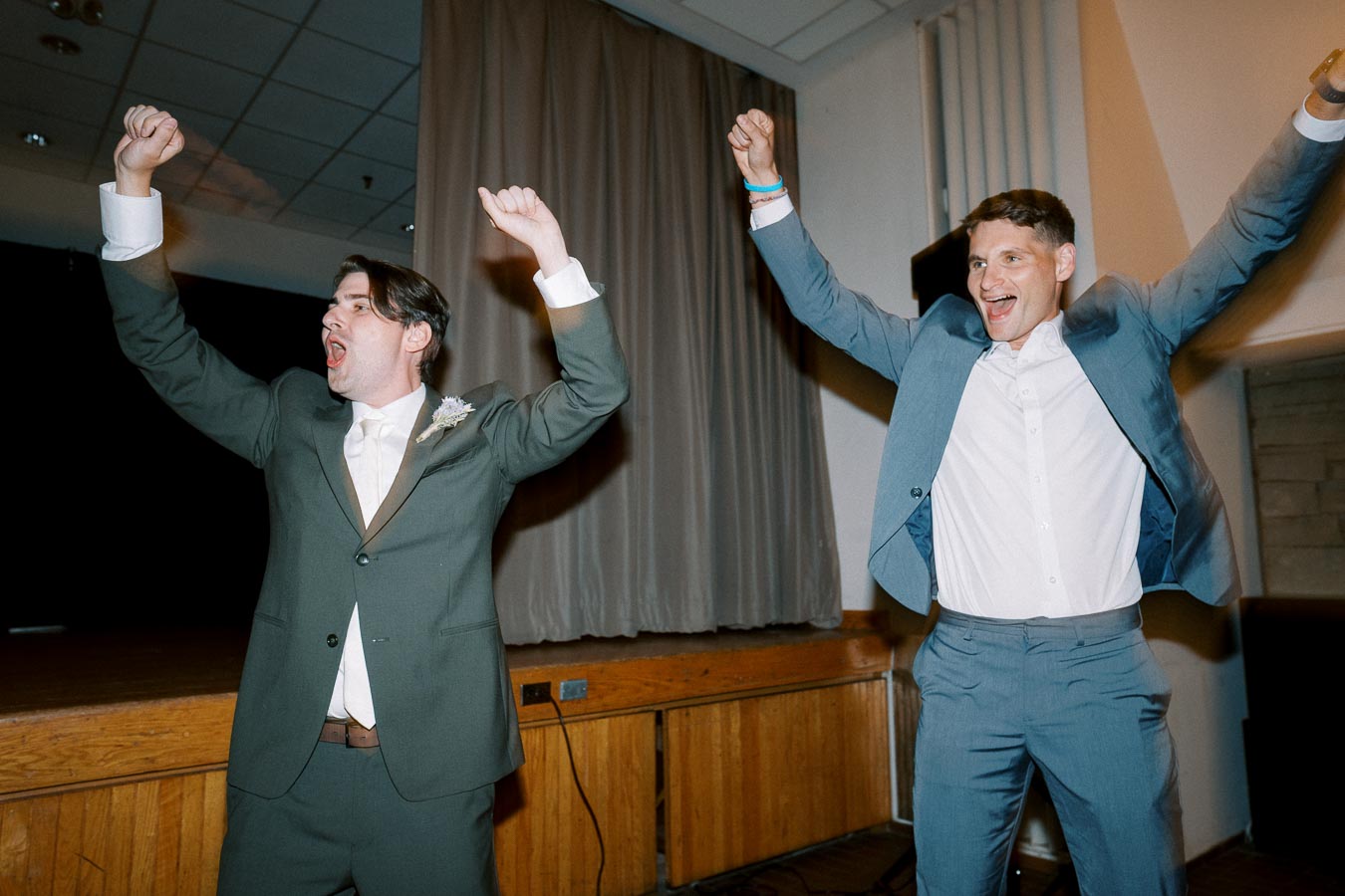 Two men celebrating enthusiastically indoors, wearing formal suits with arms raised in joy.