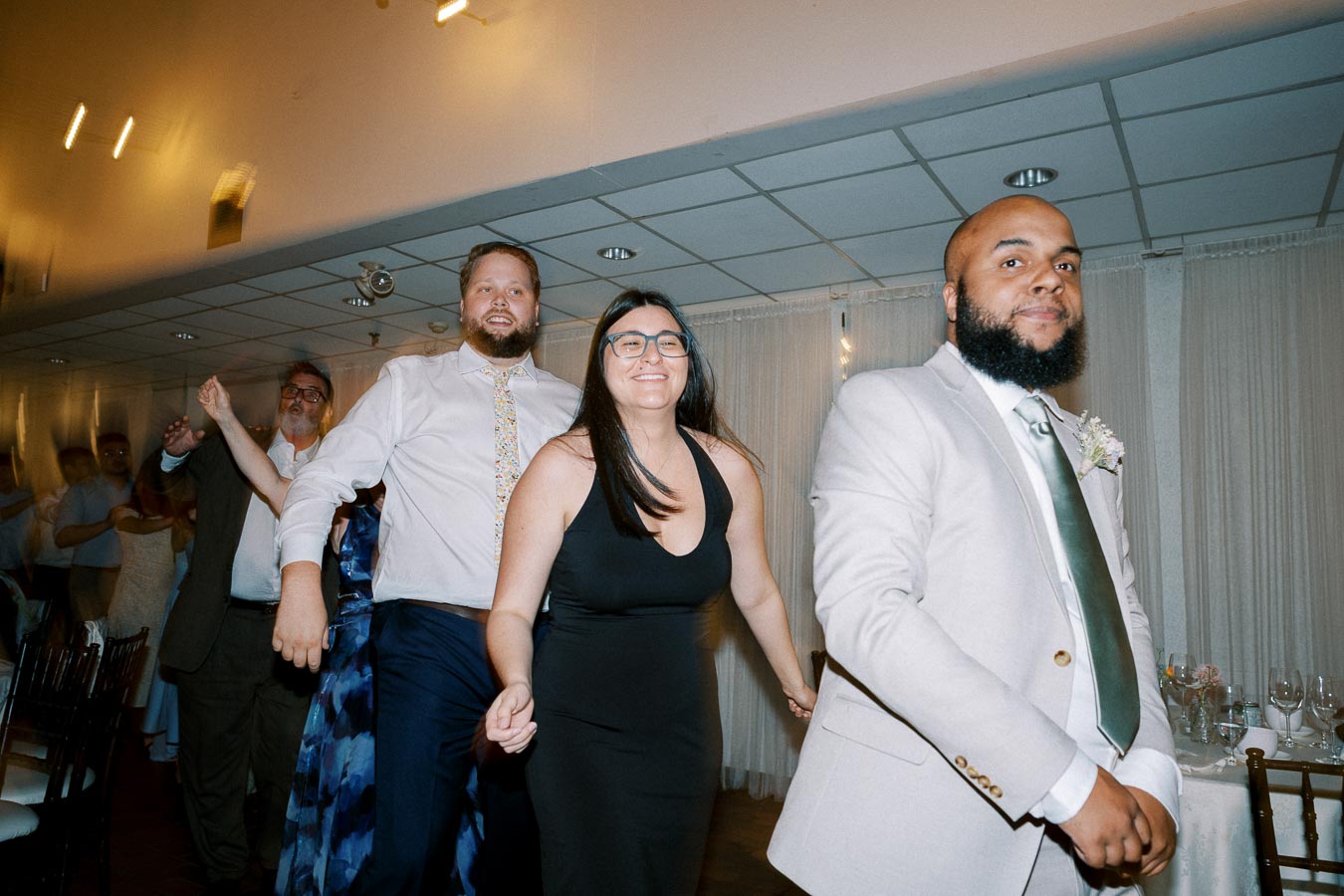 Group of people dancing in a line at an indoor event, wearing formal attire, with a joyful expression in a reception hall.