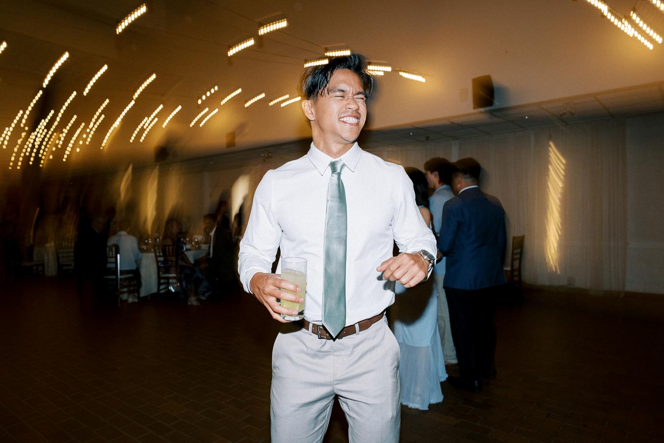A person in a white shirt and tie smiling and holding a drink, dancing at an indoor event with blurred twinkling lights and people in the background, capturing a joyful moment.