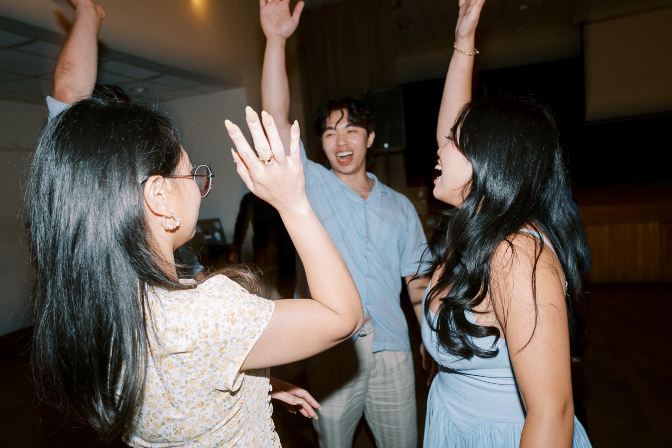 Three friends joyfully dancing together at a party, raising their hands in celebration, wearing casual summer outfits, and enjoying the lively atmosphere indoors.