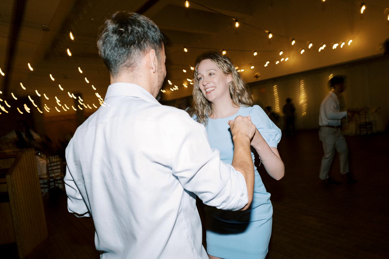 A couple dancing happily at a wedding reception, surrounded by string lights in a warmly lit venue.