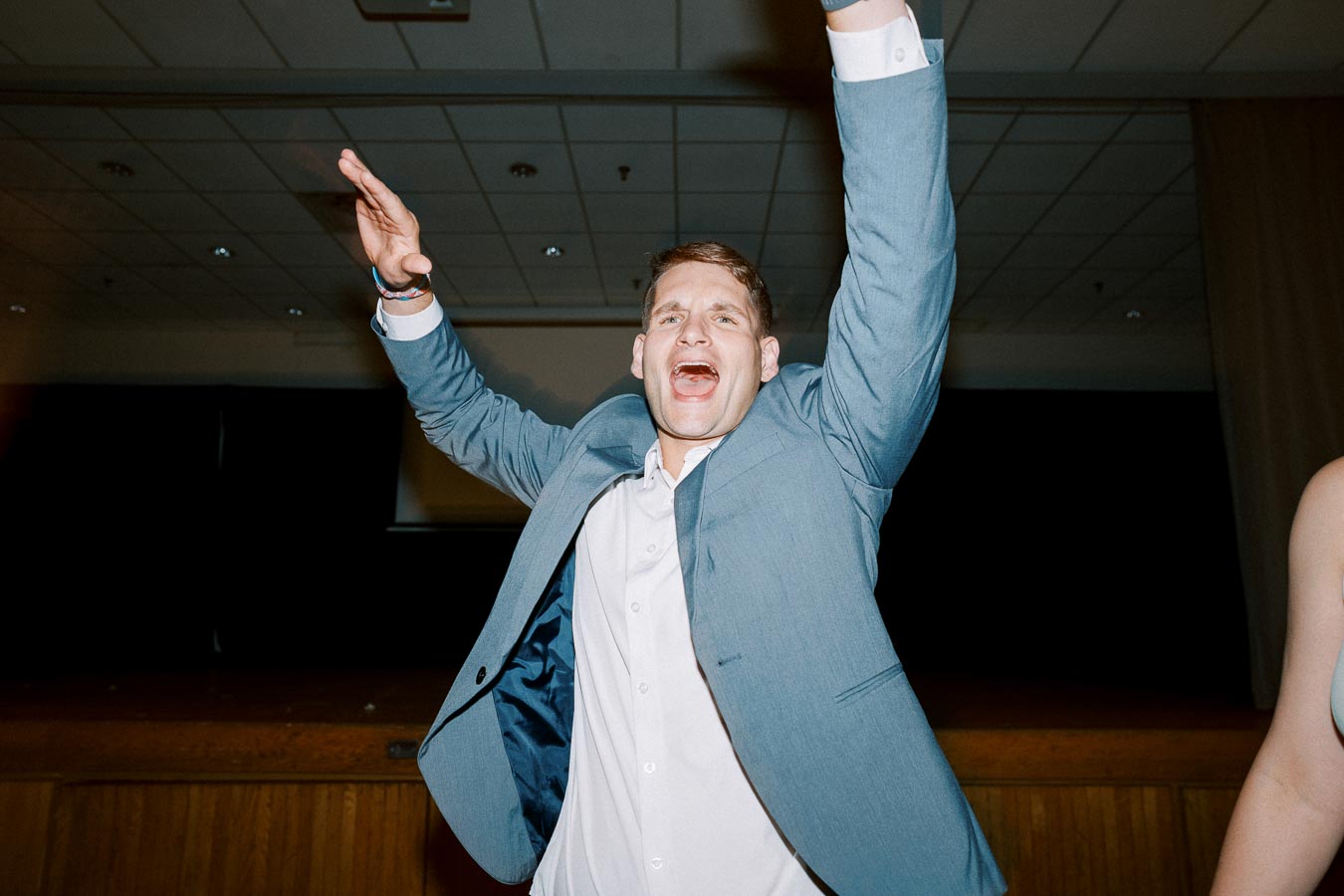 Man in a blue suit jacket enthusiastically cheering with arms raised at an event in a dimly lit venue.