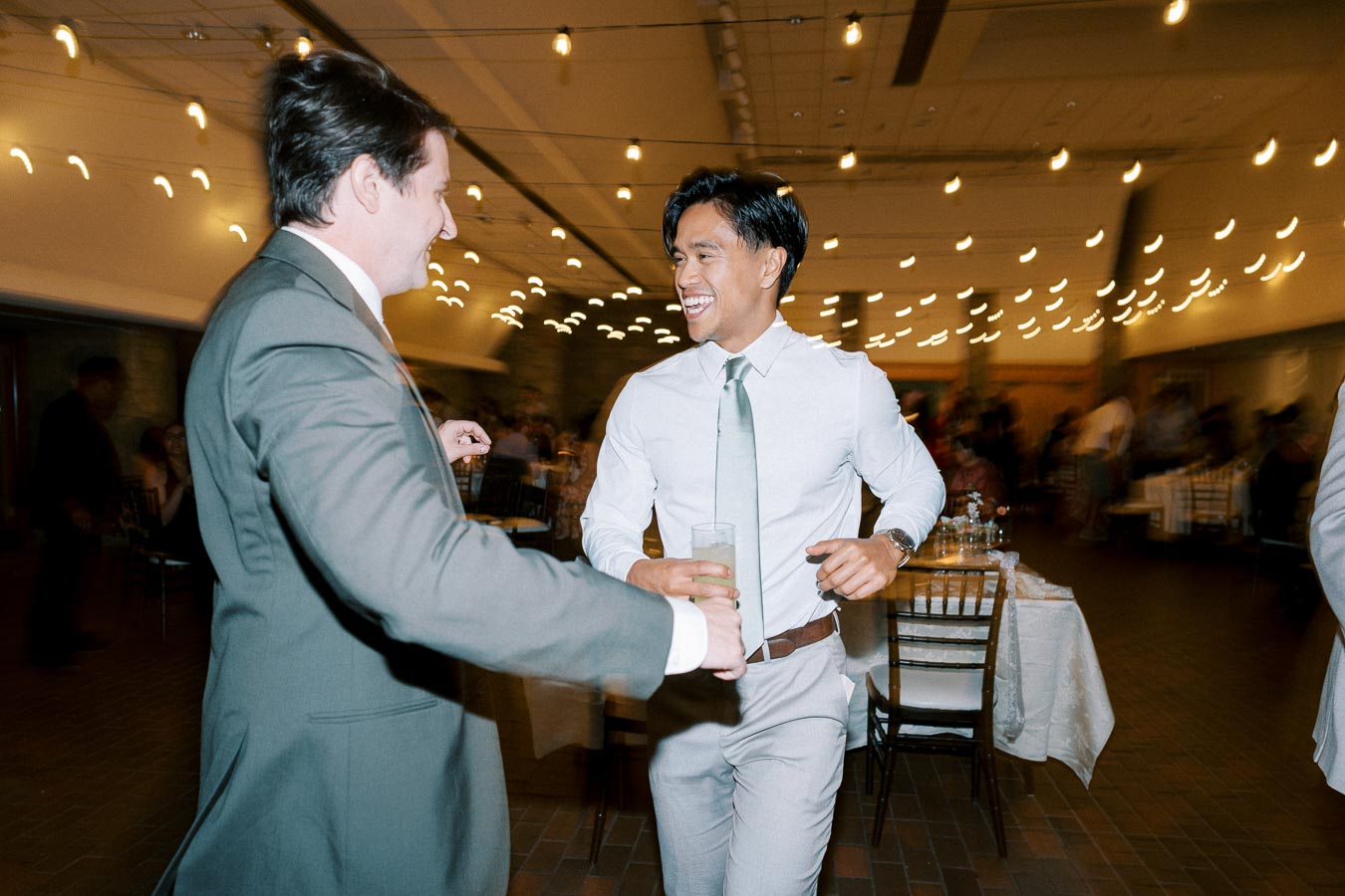 A joyous moment captured at a wedding reception with two men in suits dancing and laughing under string lights, holding drinks in their hands.