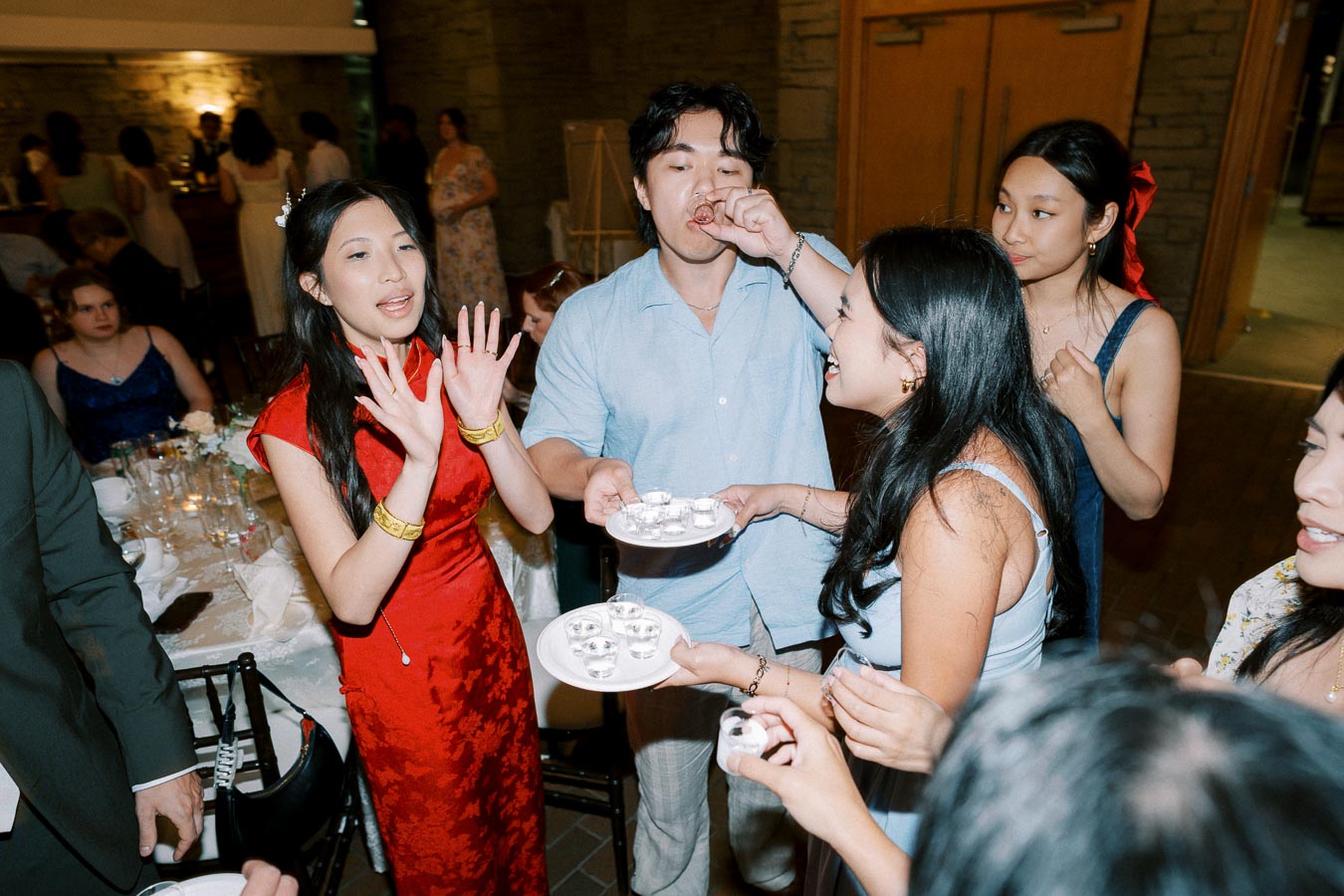 A group of people enjoy a lively gathering, with one person in a red dress talking animatedly, while another takes a shot, surrounded by friends holding trays of drinks.