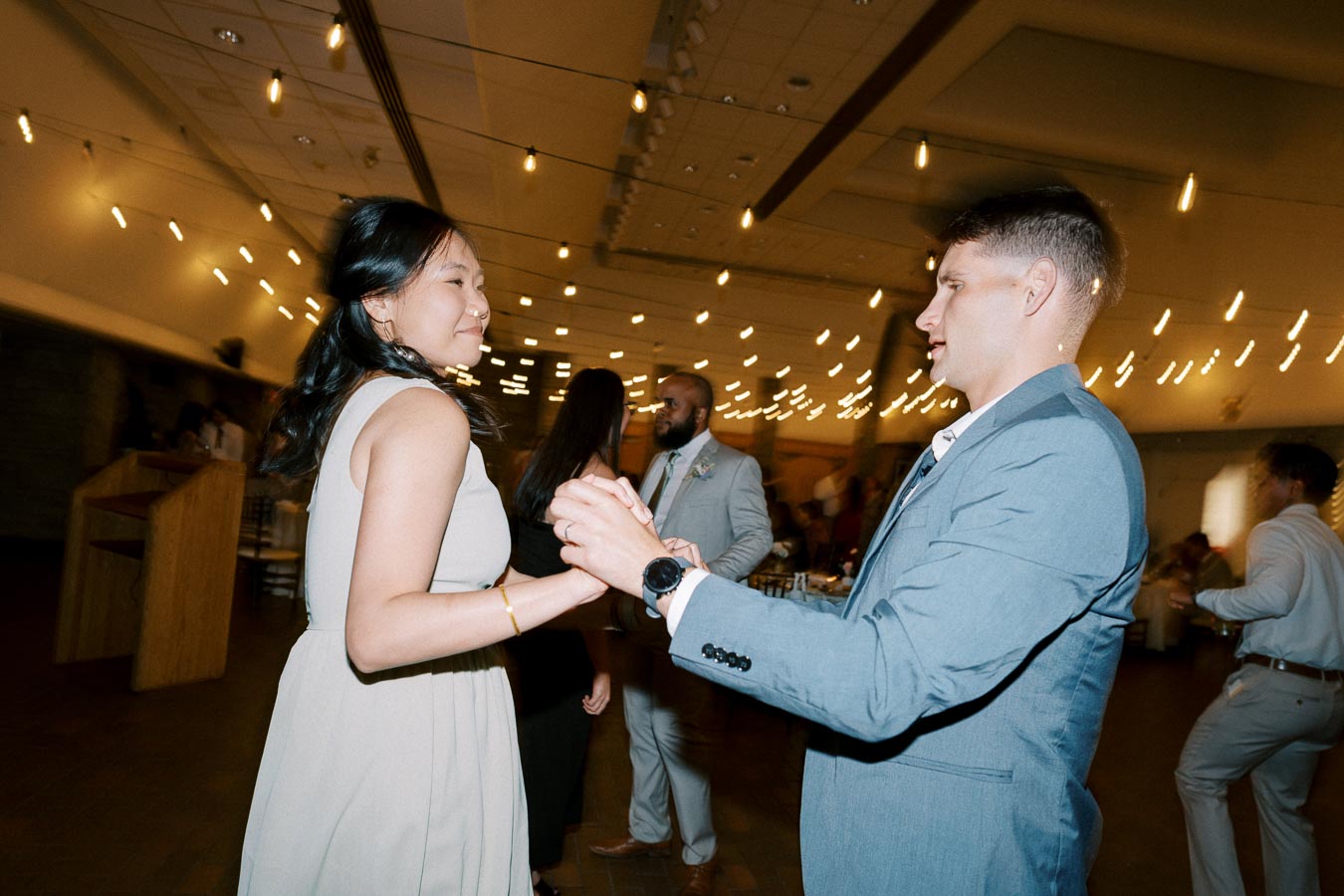 A couple dancing at a lively indoor event, surrounded by hanging string lights and other guests.