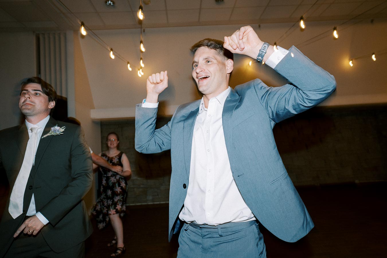 Two men joyfully dancing at a wedding reception, one wearing a light blue suit and the other in a dark suit with a floral boutonniere, under strings of warm lights.