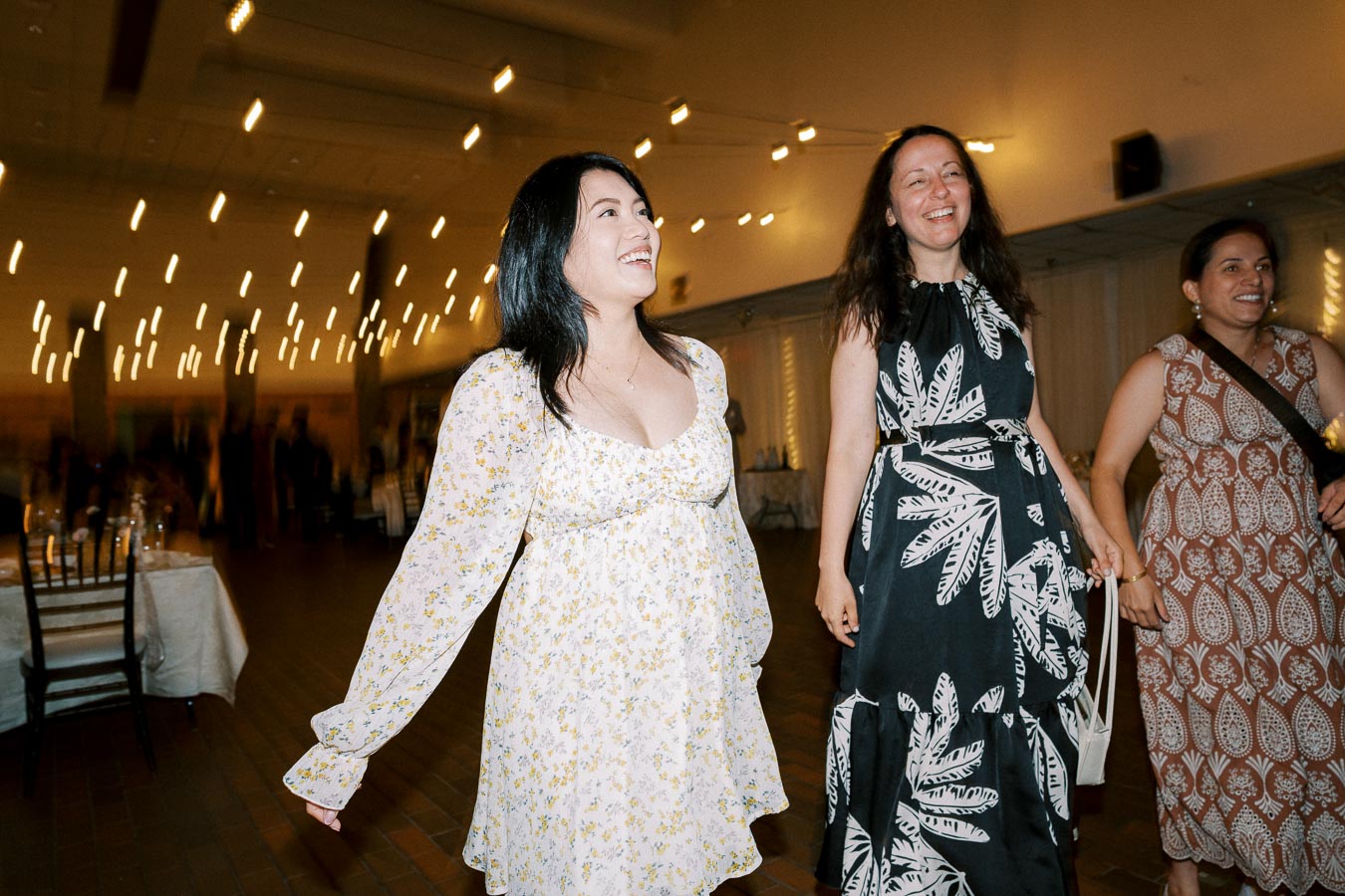 Women in floral and patterned dresses enjoying a lively indoor event with ambient lighting.