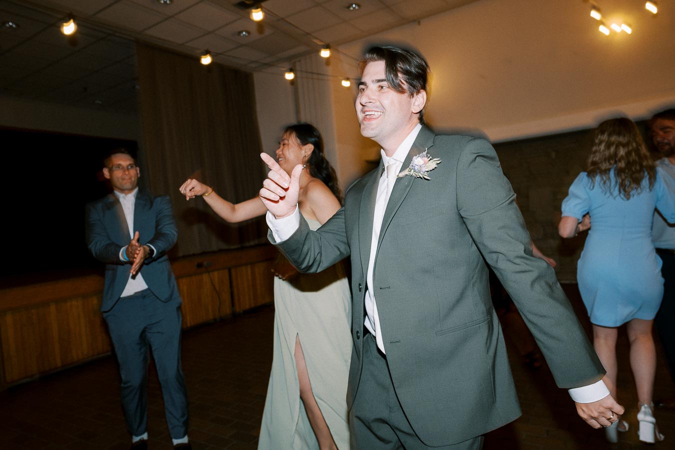 A group of people in formal attire dancing joyfully at a wedding reception, with a man in a green suit smiling and leading the dance under soft, ambient lighting.