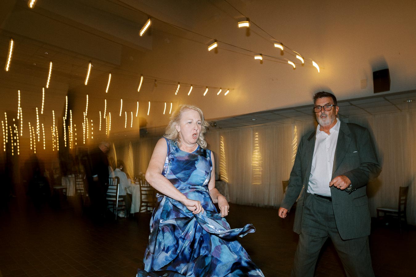 A middle-aged couple dancing energetically at a wedding reception, with blurred lights creating a dynamic background atmosphere.