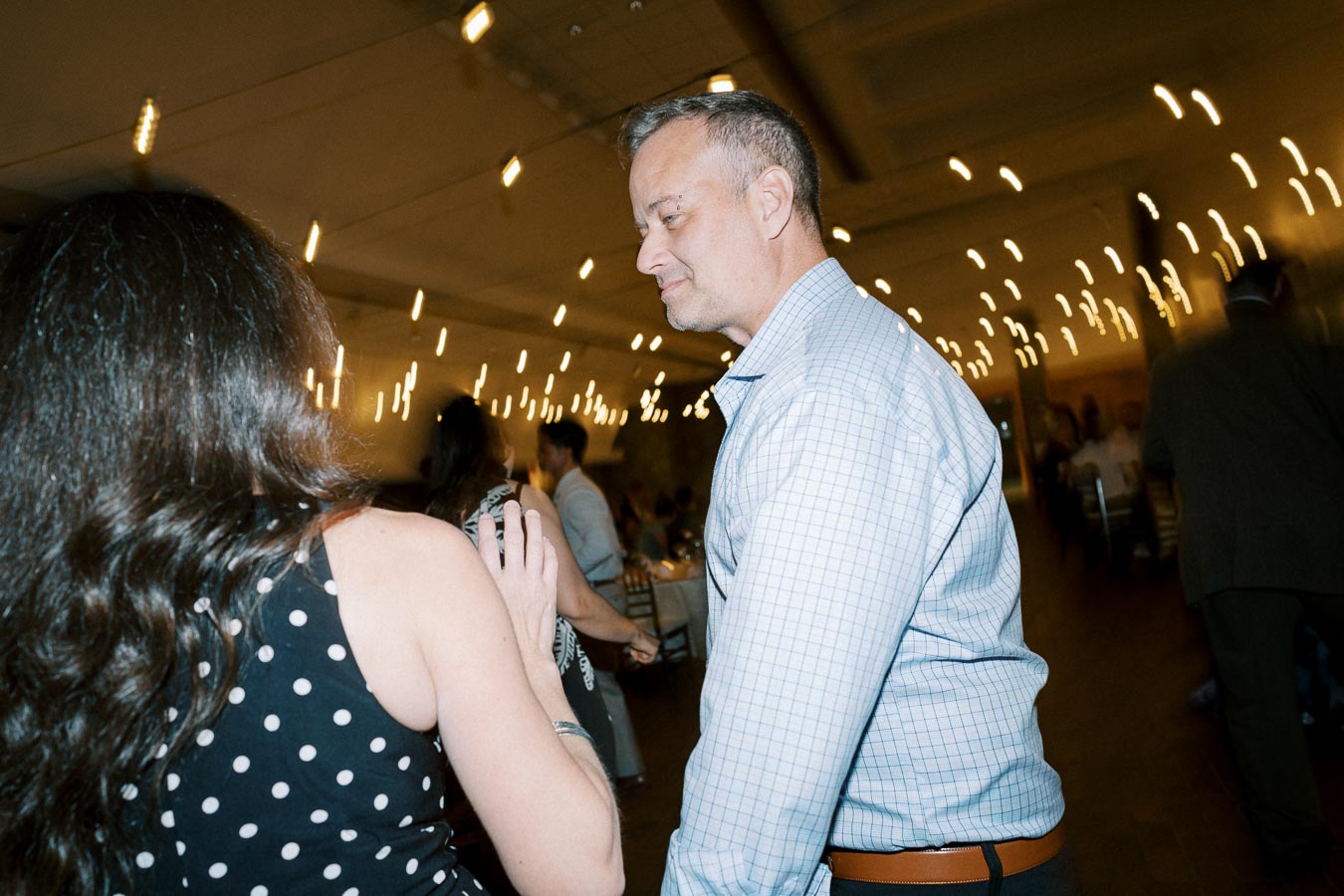 A man and a woman engage in conversation at a warmly lit indoor event, with string lights creating a festive ambiance.