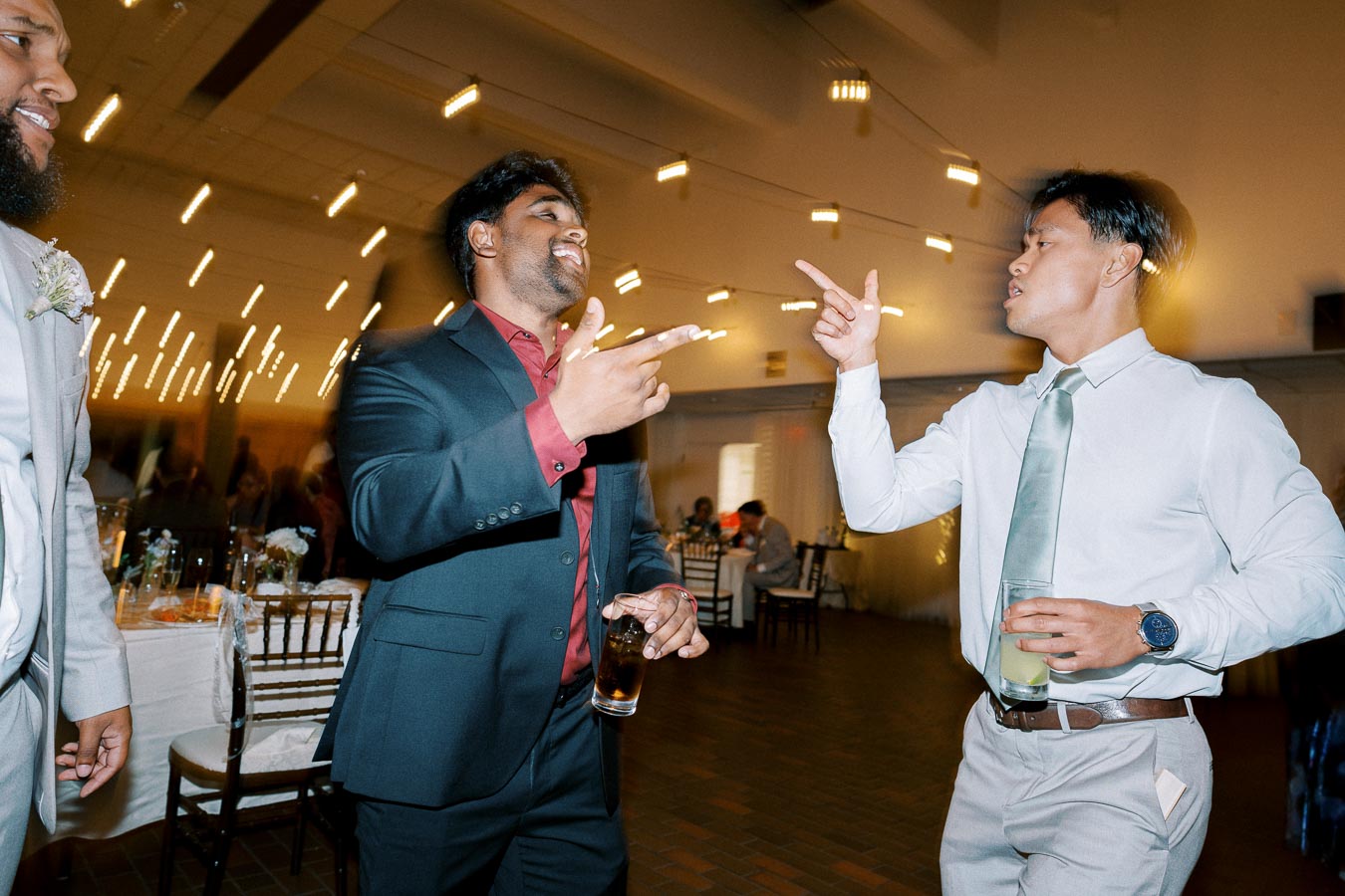 A group of men in formal attire enjoy a lively conversation and drinks at a festive indoor celebration, with string lights creating a warm ambiance.